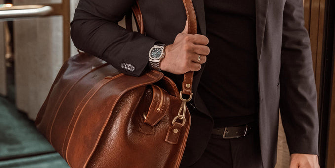 Man in a suit carrying a large brown leather weekender bag with a shoulder strap.