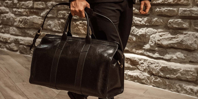 Man in a dark suit carrying a single dark brown leather weekender bag by its handles.