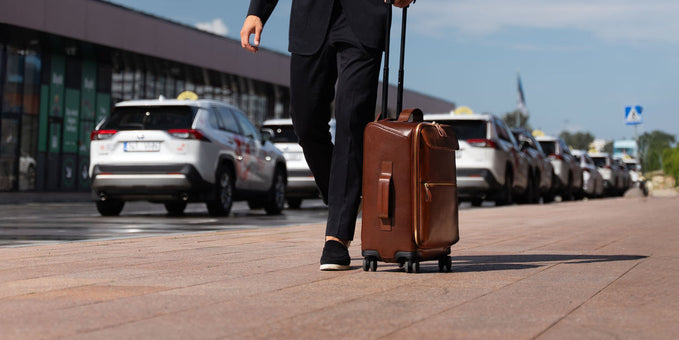 Businessman in a black suit pulling a brown leather rolling carry-on through an airport.