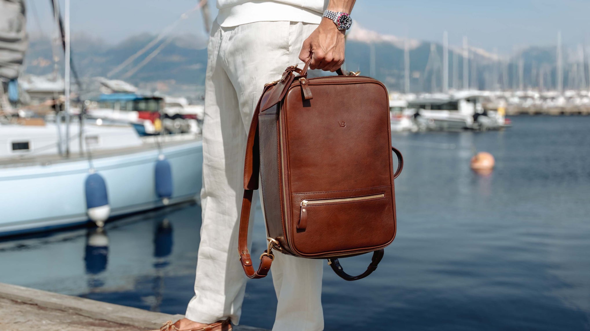 A man in white linen clothing holds a brown 10x full grain leather briefcase or laptop bag by von baer with a shoulder strap, standing by a harbor with boats and water in the background.