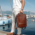 A man in a white shirt and trousers stands at a harbor holding a brown full grain leather travel backpack by von baer. The background shows calm blue water, boats, and distant mountains under a clear sky.