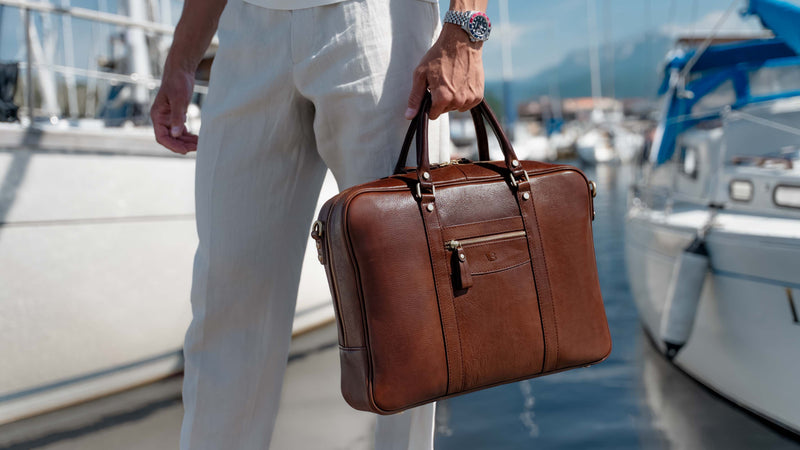 A man in white linen trousers with rolex watches carries a solid brown brown leather briefcase by Von Baer by a marina with boats in the background.