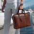 A man in white linen trousers carries a brown full grain leather briefcase by a marina with boats in the background.