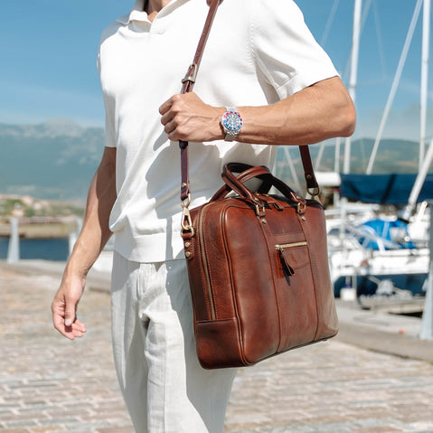 A man wearing a white polo shirt and light trousers with rolex watches carries a brown full grain leather briefcase-style shoulder bag by von baer with a long strap, standing outdoors near a marina with boats and water in the background.