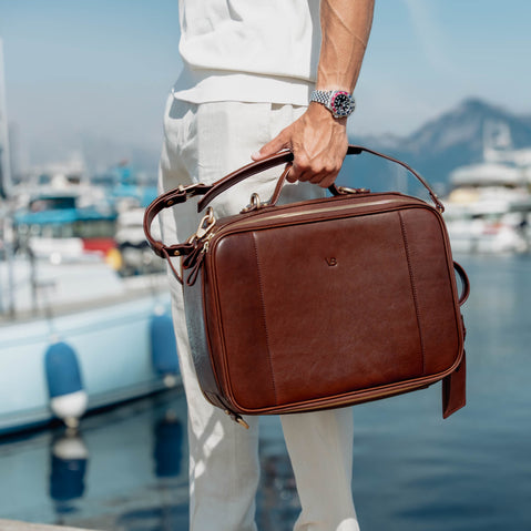 A man in a white shirt and trousers holds a brown full grain leather 2in1 briefcase/backpack by von baer with a top handle and shoulder strap, set against a blurred marina background with boats and water.