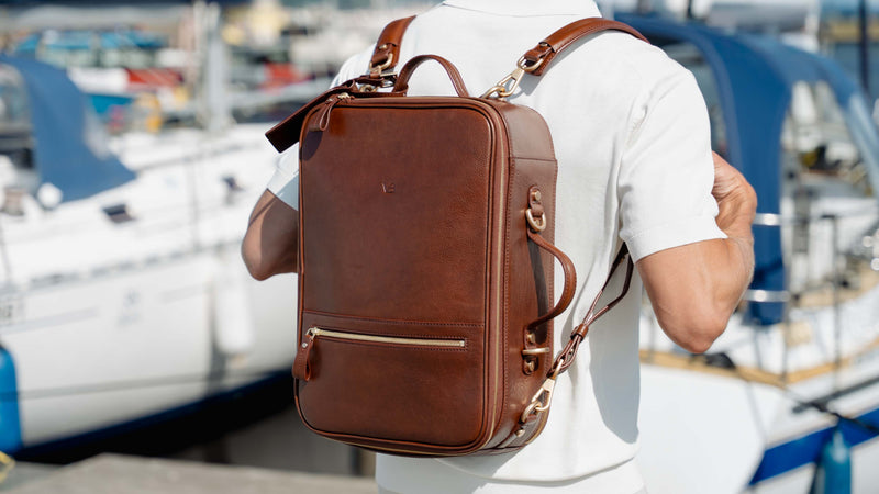 A man wearing a white t-shirt is shown from behind, carrying a brown leather messenger backpack by von baer for laptop with gold-tone hardware in a sunny outdoor setting with boats in the background.
