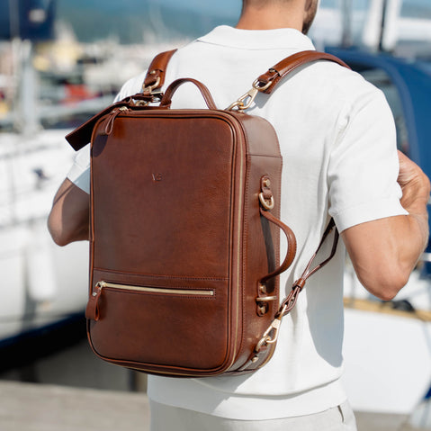 A man wearing a white polo shirt is shown from behind, carrying a brown full grain leather travel backpack by von baer with gold-tone hardware on a sunny day by the water.