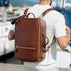 A man wearing a white polo shirt is shown from behind, carrying a brown full grain leather travel backpack by von baer with gold-tone hardware on a sunny day by the water.
