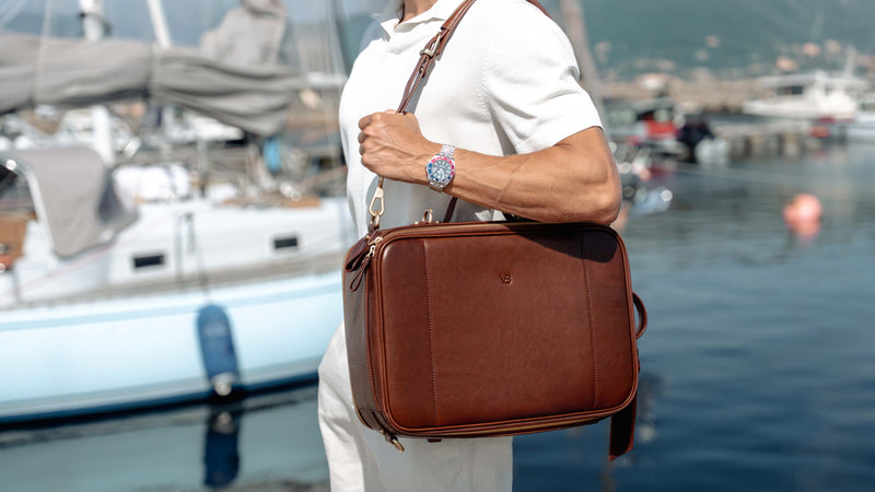 A man in a white shirt carrying a brown full grain leather briefcase/backpack/satchel bag by von baer with a shoulder strap, standing near a harbor with boats and water in the background.