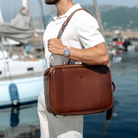A man in a white polo shirt with rolex watches carries a brown full grain leather briefcase-style shoulder bag with gold-tone hardware near a waterfront marina with boats and water in the background.