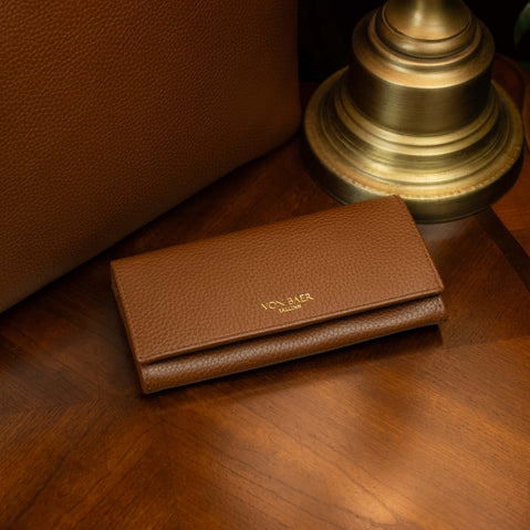 Von Baer Allure large brown full-grain leather accordion wallet displayed on a vintage wooden table next to a lamp and a luxury tote bag.