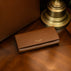 Von Baer Allure large brown full-grain leather accordion wallet displayed on a vintage wooden table next to a lamp and a luxury tote bag.
