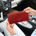 Von Baer Allure large red pebbled leather accordion wallet with gold-tone branding, held in a front view against a blurred Mercedes car interior background.