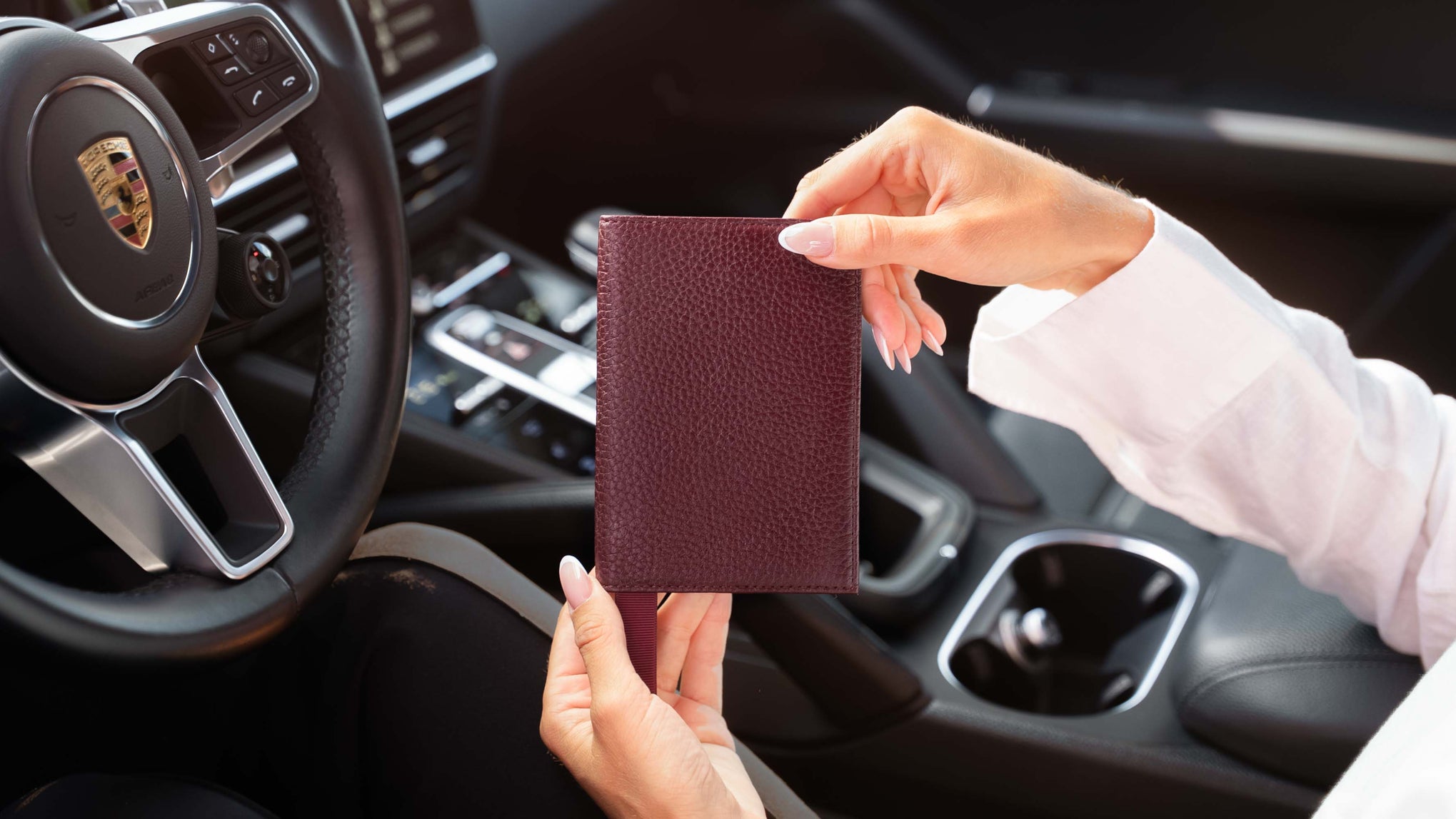 A woman holding a burgundy textured leather passport holder inside a car, with a Porsche steering wheel visible in the background.