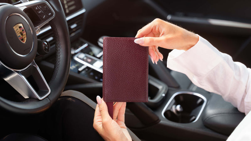A woman holding a burgundy textured leather passport holder inside a car, with a Porsche steering wheel visible in the background.