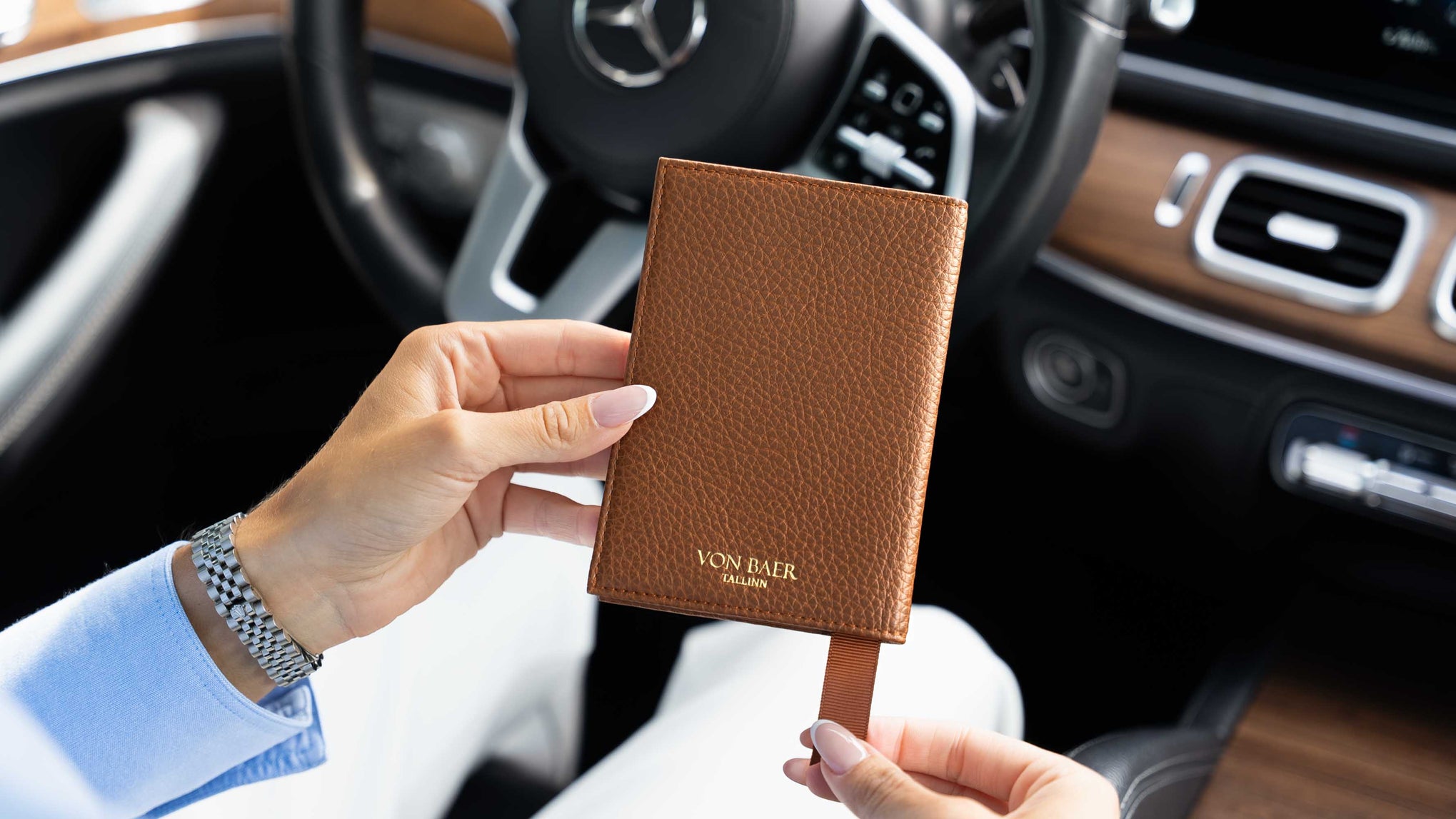 A woman holding a brown textured leather passport holder with a matching strap in front of a car interior, featuring the gold embossed brand name 'VON BAER'.