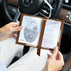 A person holds an open beautiful brown leather passport holder by Von Baer inside a car, showing US passport and a brown ribbon bookmark. The Mercedes car interior is visible in the background.