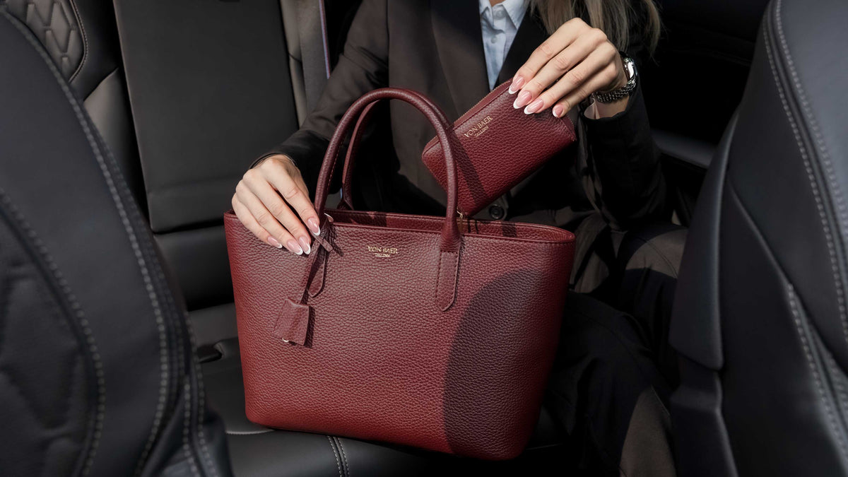Von Baer Celeste burgundy compact luxury leather zip-around wallet shown alongside an Estelle small leather tote bag, held by a person in a black leather car interior for an everyday carry view.