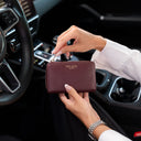 Von Baer Celeste small burgundy full-grain leather zip-around wallet held by a person in a white dress shirt inside a luxury Porsche car interior, featuring gold-tone hardware and gold-embossed Von Baer Tallinn branding.