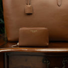 Von Baer Celeste small luxury compact tan leather zip-around wallet, displayed on a vintage wooden table next to a leather tote bag.