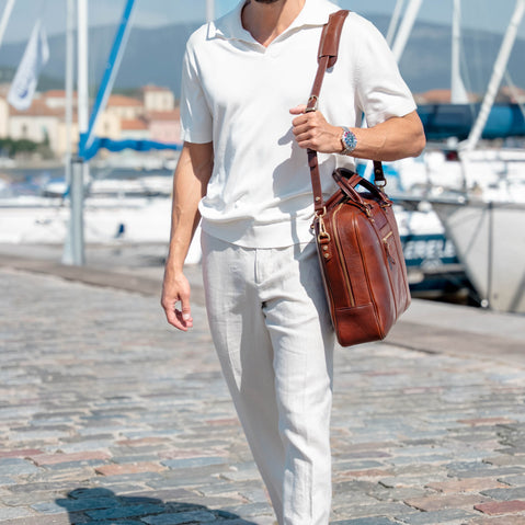 A man wearing a white short-sleeve polo shirt and white trousers walks along a cobblestone harbor path while carrying a large solid brown full grain leather shoulder bag.