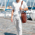 A man wearing a white short-sleeve polo shirt and white trousers walks along a cobblestone harbor path while carrying a large solid brown full grain leather shoulder bag.