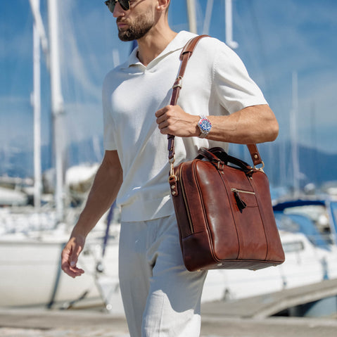 A man in a white polo shirt and trousers walks along a marina path carrying a solid brown full grain leather briefcase-style shoulder bag with a long strap.