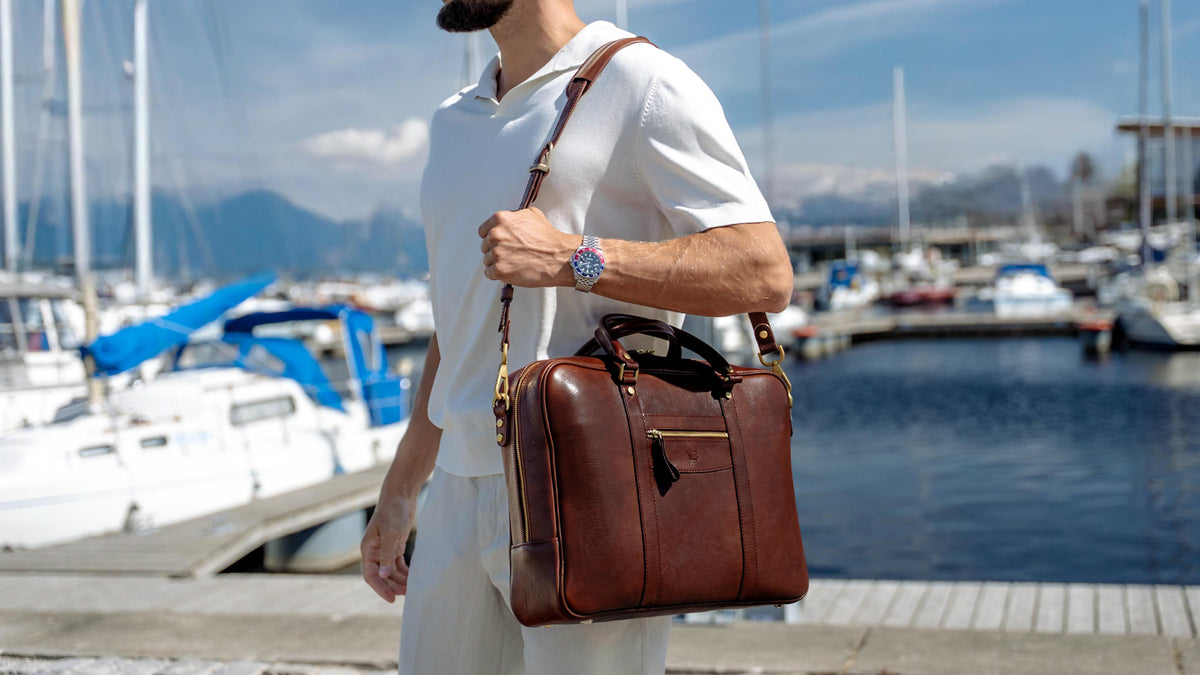A man in a white polo shirt and white pants carries a brown full grain leather briefcase bag by von baer over his shoulder while standing at a marina with boats and water in the background.