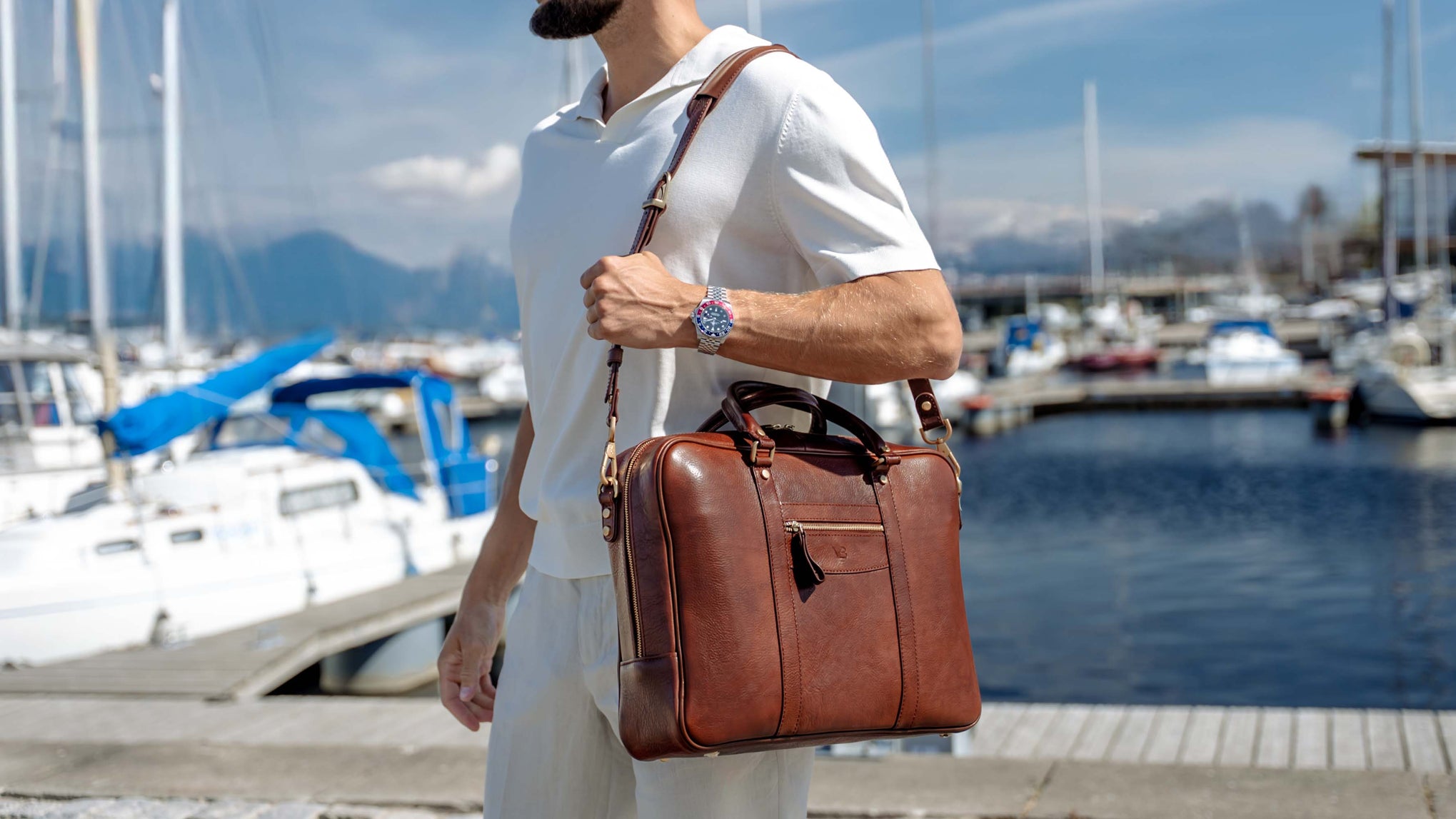 A man in a white polo shirt and white pants carries a brown full grain leather briefcase bag by von baer over his shoulder while standing at a marina with boats and water in the background.