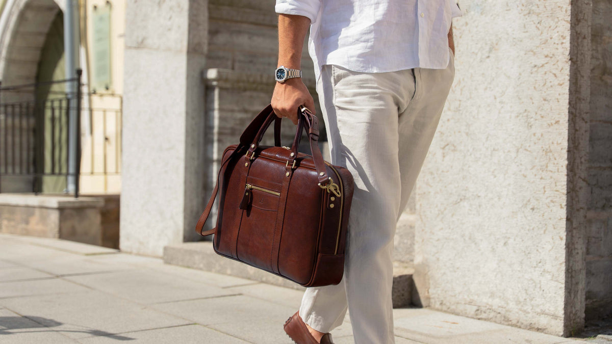 A man in a white shirt and light beige trousers carries a brown leather briefcase or duffel bag with gold-tone hardware while walking outdoors against a stone building.