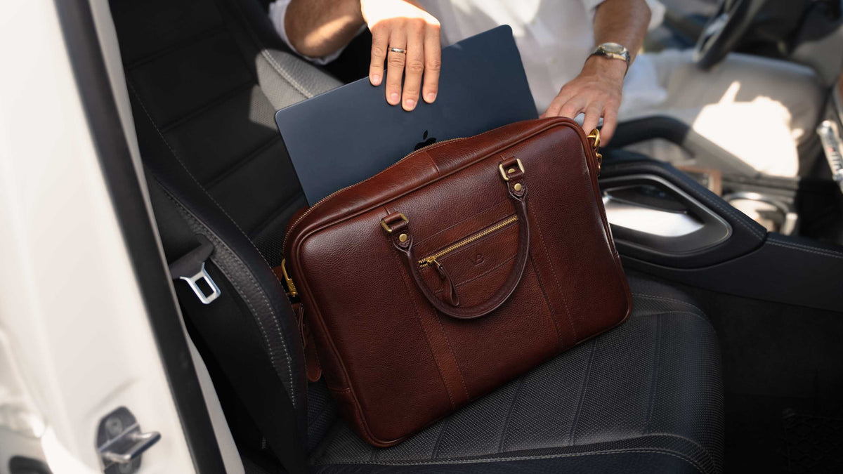A brown leather briefcase with gold-tone hardware resting on a black car seat, with a person's hands holding a dark laptop above it.