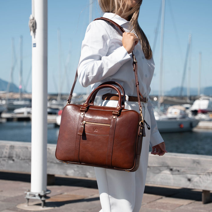 A woman in a white outfit carries a brown von baer leather briefcase-style shoulder bag with top handles and a long strap, standing outdoors near a marina with boats and water in the background.