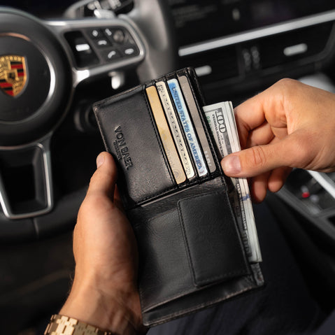 Von Baer Classic bifold leather wallet in black, stuffed with cash and credit cards like American Express, shown with a man taking out dollars in a Porsche interior