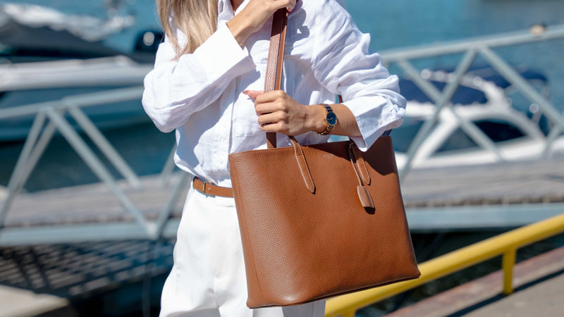 A woman in a white shirt and white pants carries a large luxury von baer tan leather tote bag with double shoulder straps in an outdoor harbor setting.