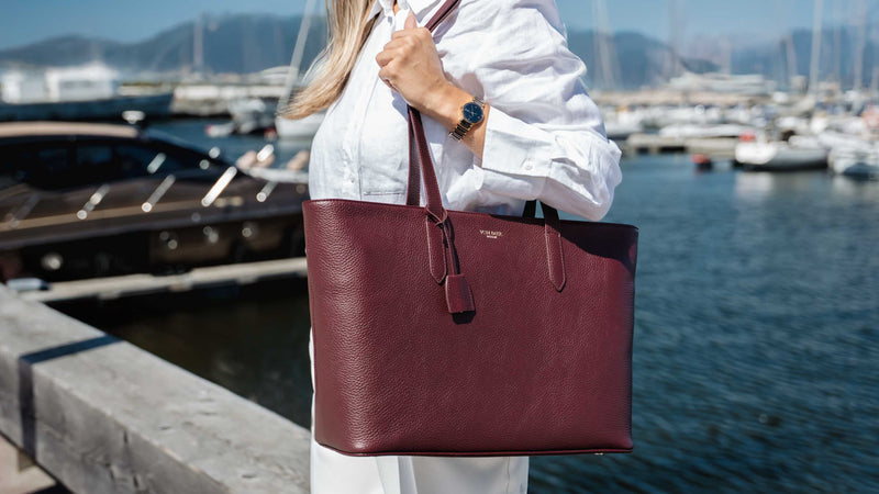 A woman in a white shirt carries a large burgundy leather tote bag by Von Baer with a tassel detail, set against a marina background with boats and water.