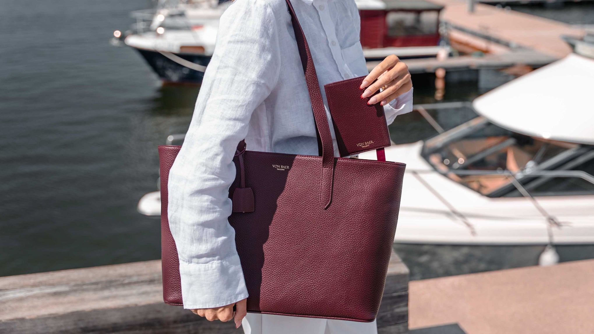 A person at a harbor carrying a luxury burgundy leather tote bag and matching passport cover. The woman is wearing a white linen shirt, and boats are visible in the background.
