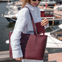 A woman at a marina carries a burgundy pebbled leather tote bag with matching wallet and matching elegant tote bag. The brand name 'Von Baer' is visible on the bag and wallet.