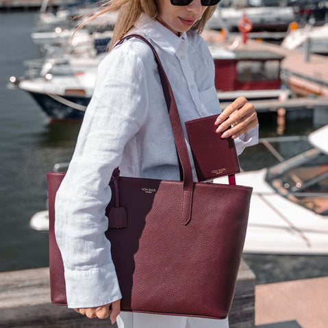 A woman at a marina carries a burgundy pebbled leather tote bag with matching wallet and matching elegant tote bag. The brand name 'Von Baer' is visible on the bag and wallet.