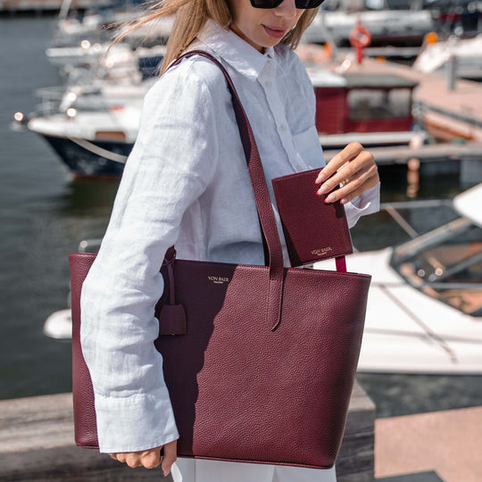 A woman at a marina carries a burgundy pebbled leather tote bag with matching wallet and matching elegant tote bag. The brand name 'Von Baer' is visible on the bag and wallet.