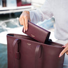 A burgundy pebbled leather tote bag for women with matching wallet, shown in a lifestyle setting with a person's hands holding it near a waterfront.