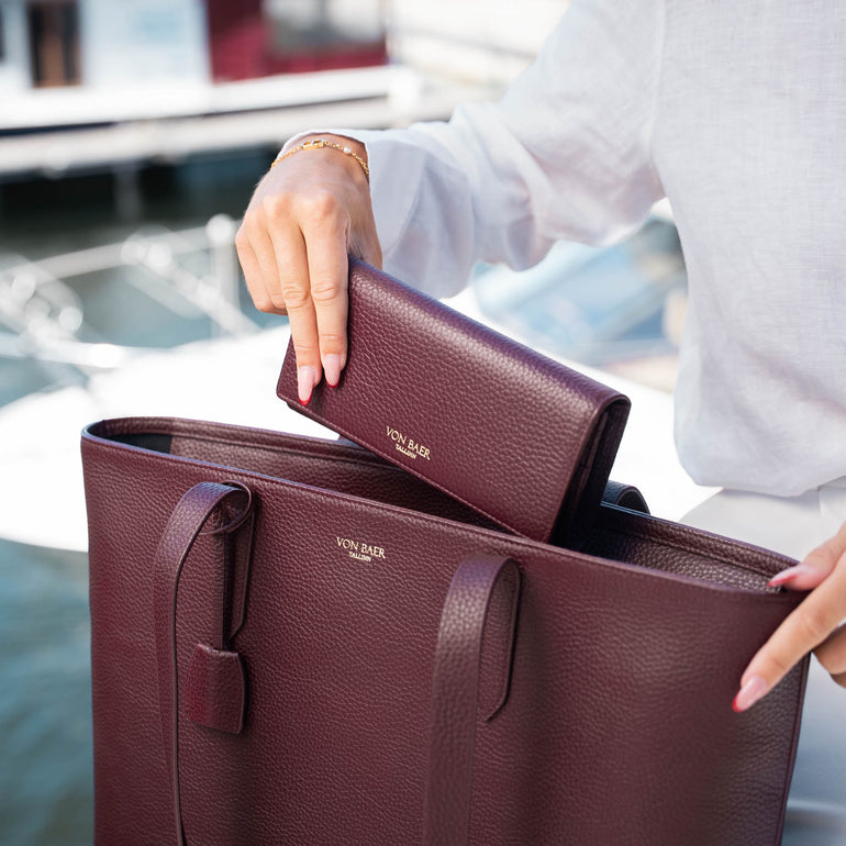 A burgundy pebbled leather tote bag for women with matching wallet, shown in a lifestyle setting with a person's hands holding it near a waterfront.
