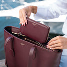 A burgundy pebbled leather tote bag with matching zip-around wallet, shown being opened by a person in a white top with water and a boat in the background. The brand name 'Von Baer' is visible in gold on both the bag and wallet.