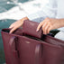 A burgundy pebbled leather tote bag with a matching zip-around wallet being held open by hands, set against a blurred outdoor background with water and a boat.