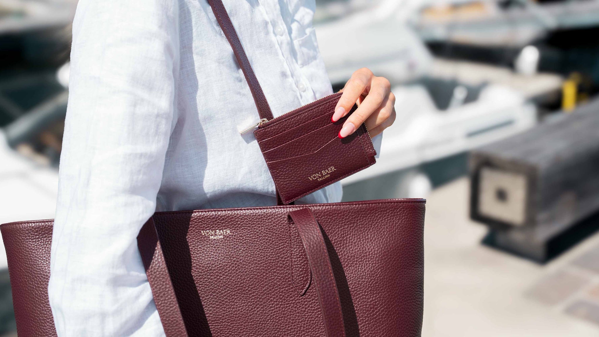 A person in a white shirt carries a luxury large burgundy pebbled leather tote bag and holds a matching small card holder against a blurred outdoor background.