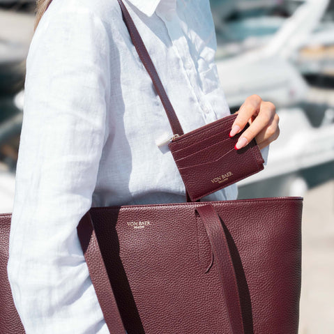 A women in a white linen shirt carries a large luxurious burgundy pebbled leather tote bag and holds a matching burgundy card holder with gold-tone lettering that reads 'VON EBER'.