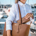 Von Baer Elegance classic tan pebbled leather tote bag with a matching Allure large leather accordion wallet tucked inside, held by a woman in a white linen shirt against a blurred marina background.
