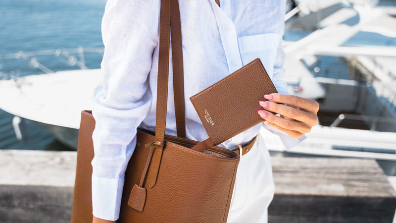 A woman in a white shirt carries a large brown leather tote bag and holds a matching brown leather passport cover with the brand name 'Von Baer' visible on the front, set against a blurred background of water and boats.