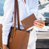 A person wearing a white linen shirt carries a large tan pebbled leather shoulder bag and holds a matching brown leather passport holder with the brand name 'Von Baer' embossed on it, set against a blurred background of water and boats.
