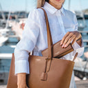 A woman in a white linen shirt carries a large brown pebbled leather tote bag with a matching zip-around wallet tucked into the bag's opening, set against a blurred marina background.