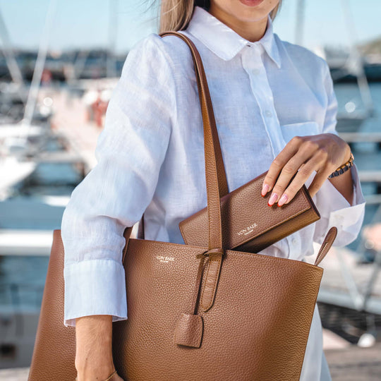A woman in a white linen shirt carries a large brown pebbled leather tote bag with a matching zip-around wallet tucked into the bag's opening, set against a blurred marina background.